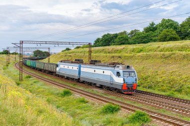 A powerful gray electric locomotive pulls a long train of wagons loaded with coal to the railway station in the seaport. Spring.