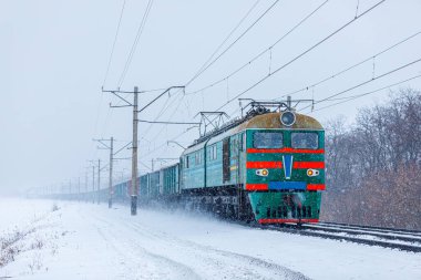 A powerful dual-system electric locomotive pulls a long freight train to the railway station. Snowstorm. Intermodal freight railway transportation of goods.