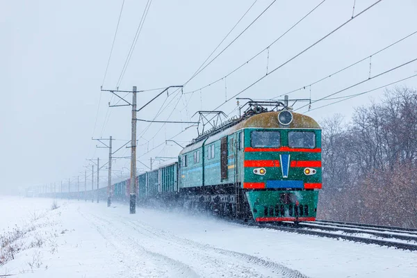Güçlü bir çift sistem elektrikli lokomotif uzun bir yük trenini tren istasyonuna çeker. Kar fırtınası. Malların intermodal yük taşımacılığı.