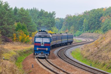 Mavi bir lokomotif viraj yolu boyunca bir yolcu trenini tren istasyonuna çeker. Yeşil ve sarı yapraklı ağaçlar. Akşam sonbahar aydınlatması.