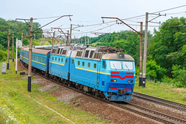  Güçlü iki bölümlü mavi bir lokomotif uzun bir yolcu trenini tren istasyonuna çeker. Yeşil yapraklı ağaçlar. Akşam ışığı..