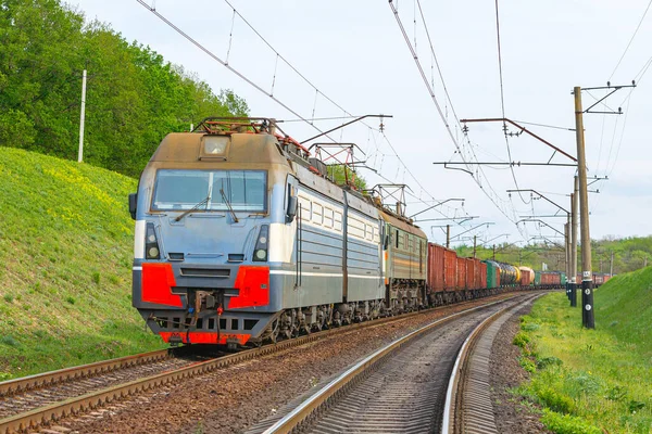 Güçlü yeşil bir elektrikli lokomotif demiryolu istasyonundan ürünlerle dolu uzun bir treni çeker. Günbatımı bahar ışığı.