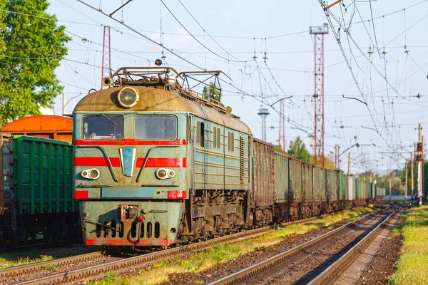 Güçlü yeşil bir elektrikli lokomotif demiryolu istasyonundan ürünlerle dolu uzun bir treni çeker. Günbatımı bahar ışığı.