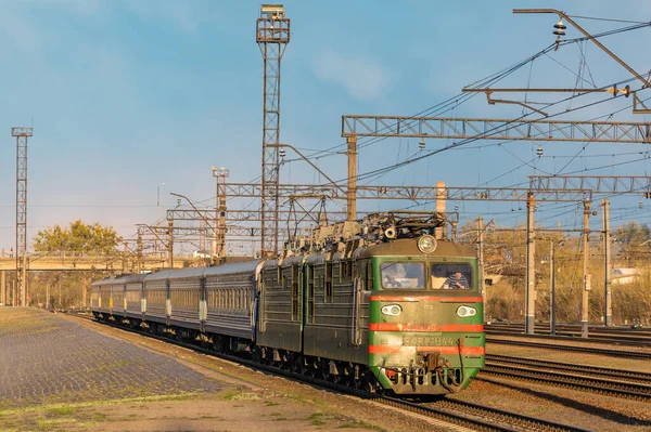 Kharkiv, Ukrayna - Nisan 2021: Yeşil bir VL82 elektrikli lokomotif, yağmurlu bir gökyüzünün arka planında batan güneşin ışınlarıyla yolcu trenini tren istasyonuna çekiyor. Ukrayna demiryolları