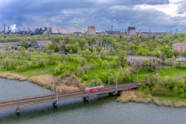 Kryvyi Rih, Ukrayna - 2021 İlkbaharı: Kırmızı bir Tatra T3 tramvayı, büyük bir metal fabrikasının arka planına karşı bir göletin üzerinden geçiyor. Yukarıdan görüntüle.