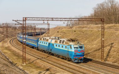 Mavi elektrikli bir lokomotif Chs7, tren rayları boyunca şehirlerarası uzun bir yolcu trenini çekiyor. Kış güneşli hava.