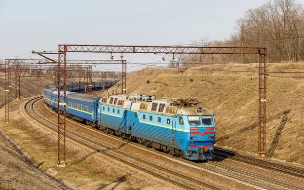 Mavi elektrikli bir lokomotif Chs7, tren rayları boyunca şehirlerarası uzun bir yolcu trenini çekiyor. Kış güneşli hava.