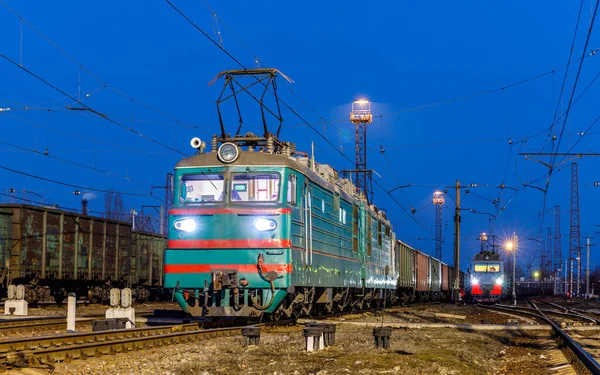 İki elektrikli lokomotif yük trenleriyle büyük bir tren istasyonunun raylarında duruyor. Ukrayna Demiryolu Akşam fotoğrafı.