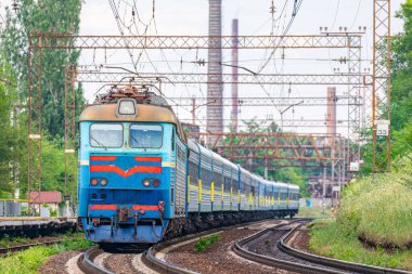 A powerful locomotive pulls a long train of blue passenger cars to the railway station. Passenger railway transportation. Close-up view. Industrial landscape in the background.