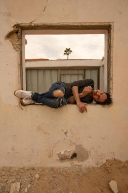 Bir adam terk edilmiş ve yıkılmış bir evin boş penceresinde yatıyor.