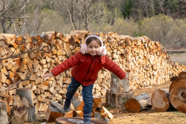 Little girl playing on wooden logs, standing on the background of wall of firewood. Country life — Stok Foto