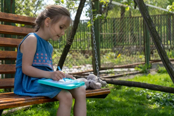 Beautiful little girl is learning to draw and write while sitting on a swing on a sunny day. — Stok Foto