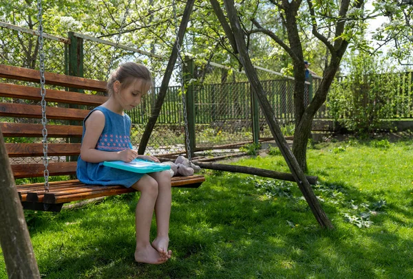 Adorable little girl is concentrated on learning to write while sitting on a swing on a sunny day — Stok Foto