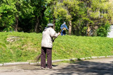 Şehirde caddeyi süpüren yaşlı bir kadın. Arka plandaki adam çimleri biçiyor.