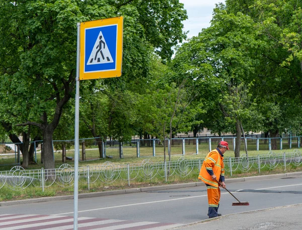 Man cleaner sweeping street with broom, municipal worker in uniform with broomstick brush in hands, cleaning service. Male janitor with broom , cleaning city from garbage near a pedestrian crossing — Stock Photo, Image
