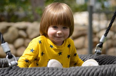 Hamak yuvasında salıncakta sallanan sevimli küçük bir çocuk. Uzun sarı saçlı, yakından çekilmiş portresi olan çocuk. Bahçede sarı tişörtlü bir çocuk..