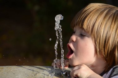 Çeşmeden su içen tatlı bir çocuk. Yaz susuzluğu. Uzun saçlı bir bebek ağzıyla bir su püskürtüsü yakalar. Oyun parkındaki çeşme.