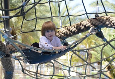 Ağlara tırmanırken tezahürat yapan mutlu şirin çocuk. Çocuk oyun sahasında halat örgüsü üzerinde sürünüyor