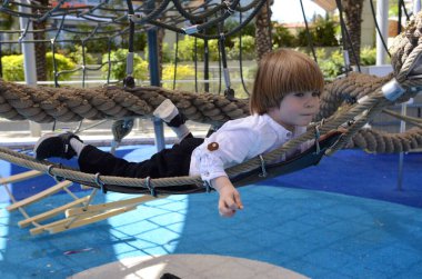 Ağlara tırmanırken tezahürat yapan mutlu şirin çocuk. Çocuk oyun sahasında ip örgüsü üzerinde sürünüyor. Beyaz gömlekli şirin bir çocuk hamakta yatıyor. Oyun parkında şebeke, ip parkı. Faaliyetler için internet. Çocuk dinleniyor.