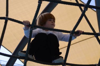 Mutlu çocuk ağa tırmanırken tezahürat yapıyor. Çocuk oyun sahasında halat örgüsü üzerinde sürünüyor