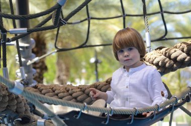 Ağlara tırmanırken tezahürat yapan mutlu şirin çocuk. Çocuk oyun sahasında ip örgüsü üzerinde sürünüyor. Beyaz gömlekli şirin bir çocuk hamakta yatıyor. Oyun parkında şebeke, ip parkı. Faaliyetler için internet. Çocuk dinleniyor.