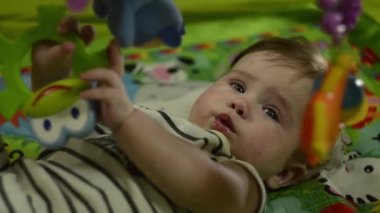 A little boy is looking at the camera. Lies on his back and plays with a mobile and soft toys. Very serious look
