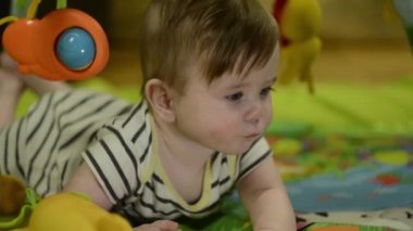 A little boy is looking at the camera. Lies on his back and plays with a mobile and soft toys. Very serious look