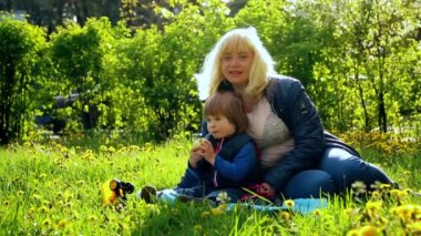 Little boy with blond hair, 2 years old, sitting with his blonde mom on the green grass, eating a banana. Nearby are a toy car and a tractor. Picnic with granny's in the city. sunlight