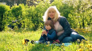 Little boy with blond hair, 2 years old, sitting with his blonde mom on the green grass, eating a banana. Nearby are a toy car and a tractor. Picnic with granny's in the city. sunlight