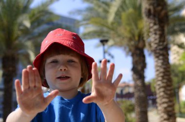 Handsome little boy in a red hat. In the background is a fountain and a palm grove. The child enjoys the summer day. Cheerful portrait. Concept: summer holidays, walk in the park.