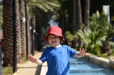 Handsome little boy in a red hat. In the background is a fountain and a palm grove. The child enjoys the summer day. Cheerful portrait. Concept: summer holidays, walk in the park.