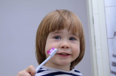 Küçük sarışın çocuk ev banyosunda dişlerini fırçalamayı öğreniyor. Çocuk nasıl sağlıklı kalacağını öğreniyor. Sağlık hizmeti konsepti. Sevimli çocuk gülümsüyor.