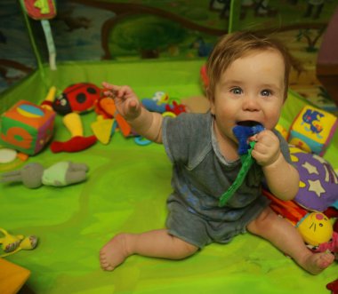 7 aylık bir çocuk yeşil bir zeminde oturuyor, gülüyor, oyuncaklarla oynuyor. Gülümseyen, kameraya bakan pozitif bir çocuk. Küçük çocuk - yeni yürümeye başlayan