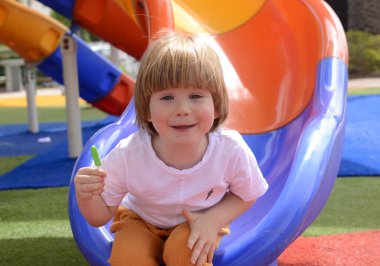 Mutlu gülümseyen bir çocuk. 3 yaşında, sarışın. Sevimli bir çocuk parkta gülüyor ve elinde yeşil bir lolipop tutuyor. Konsept: iyi ruh hali, aile tatili