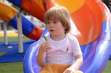 Mutlu gülümseyen bir çocuk. 3 yaşında, sarışın. Sevimli bir çocuk parkta gülüyor ve elinde yeşil bir lolipop tutuyor. Konsept: iyi ruh hali, aile tatili