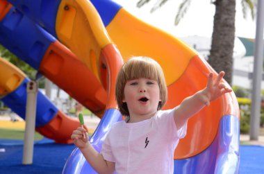 Mutlu gülümseyen bir çocuk. 3 yaşında, sarışın. Sevimli bir çocuk parkta gülüyor ve elinde yeşil bir lolipop tutuyor. Konsept: iyi ruh hali, aile tatili