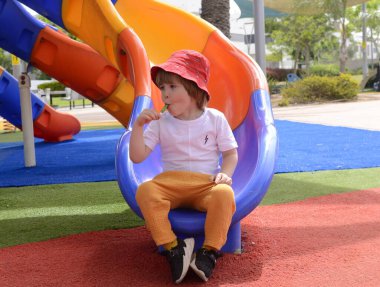 Oyun parkında yeşil lolipop yiyen küçük çocuğa yukarıdan bakın, kameraya bakıyor. Tatlıyı. Kırmızı panamalı bir çocuk. Bir çocuğun portresi.