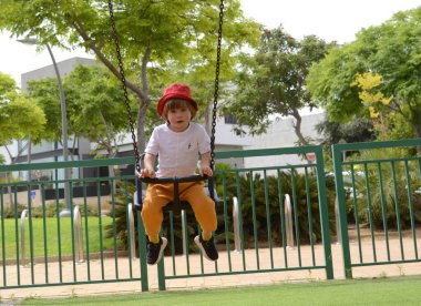 Kırmızı panama şapkalı küçük bir çocuk salıncakta sallanıyor. Oyun alanı, yaz, renkli kıyafetler.