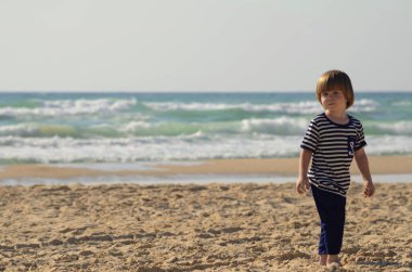 3 yaşında, uzun saçlı, çapalı, denizci çizgili bir tişörtü olan bir çocuk. Çocuk kumlu bir plajda duruyor, arka planda deniz. Aile tatili, deniz tatili, sahil tatili.