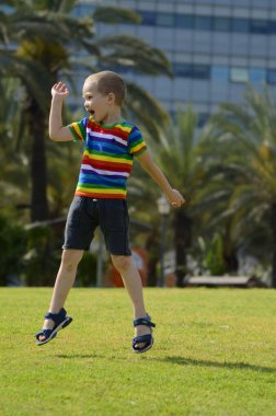 Küçük bir çocuk parktaki yeşil bir çayıra atlar. Arka planda bir Palm Avenue var. Çok renkli tişörtlü bir çocuk yazın sevinir.