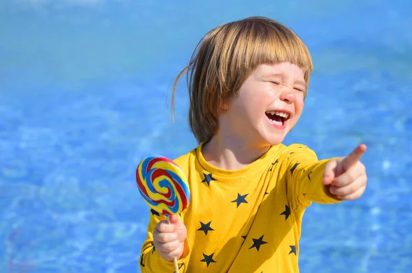 一个小男孩坐在游泳池边吃着一个色彩斑斓的棒棒糖 阳光明媚的夏天 家庭假期 与孩子一起旅行 度假胜地 水上公园 这个孩子夏天很开心 对着太阳笑 — 图库照片