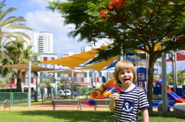 Güzel oyun alanı, çiçek açan ağaçlar, palmiye ağaçları. Sevimli küçük çocuk gülümsüyor ve çubukta şeker yiyor. Konsept, aileler için rahat bir yerleşim alanı. Anaokulu reklamı, okul.