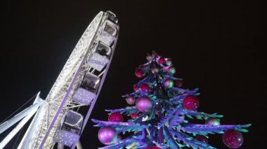 Şenlikli çelenkler içinde dönme dolap. Işıklar ve toplarla süslenmiş Noel ağacı. Noel pazarı, akşam, günün karanlık zamanları. Kavram: Noel, kış, Aralık, tatiller