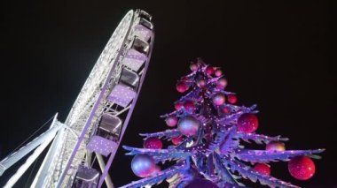 Şenlikli çelenkler içinde dönme dolap. Işıklar ve toplarla süslenmiş Noel ağacı. Noel pazarı, akşam, günün karanlık zamanları. Kavram: Noel, kış, Aralık, tatiller