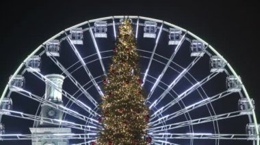 Parlak ışıklardaki Noel ağacı karanlıkta parlayan dönme dolabın arka planına karşı. Noel Fuarı. Konsept: Kış tatili, Noel pazarı, Aralık. Bokeh çelenkleri, bulanık ışık