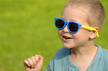 Portrait A five-year-old boy in black sunglasses against a green lawn. The child is sitting on the grass. Space for text. Summer concept: getting ready for school, a park for children, classes during the holidays, family vacation