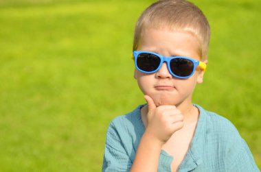 Portrait A five-year-old boy in black sunglasses against a green lawn. The child is sitting on the grass. Space for text. Summer concept: getting ready for school, a park for children, classes during the holidays, family vacation