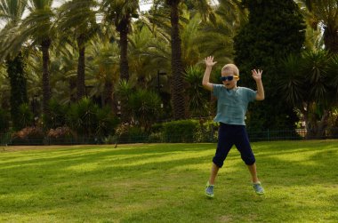The boy jumps for joy on a green field. A child in sunglasses on a background of palm trees. Concept: holidays, travel, exotic destinations, tropical countries.