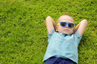 Boy schoolboy lies on a green meadow on the grass. A child with sunglasses is resting, enjoying the summer. Dreams about travel, sea, adventure, amusement park, birthday