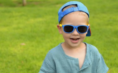 Portrait A five-year-old boy in black sunglasses against a green lawn. The child is sitting on the grass. Space for text. Summer concept: getting ready for school, a park for children, classes during the holidays, family vacation